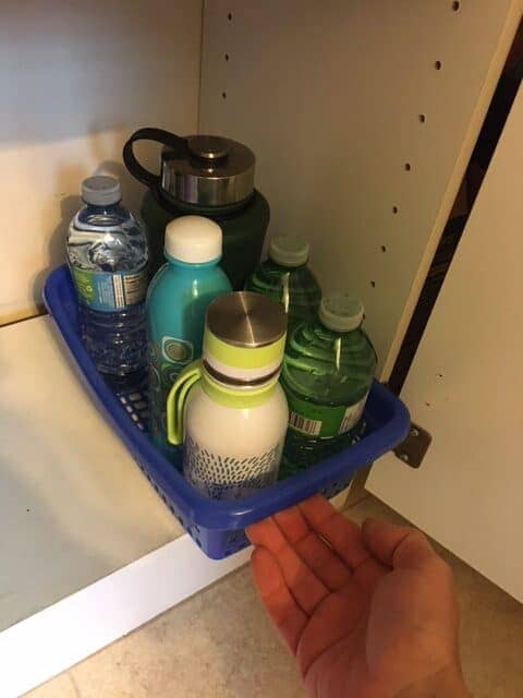 A view of a person keeping the water bottles inside a cabinet stored in a plastic basket