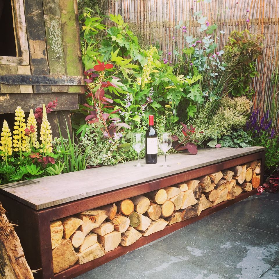 wooden bench with firewood arranged beneath it a bottle of wine and glasses placed on top of bench plants and flowers around